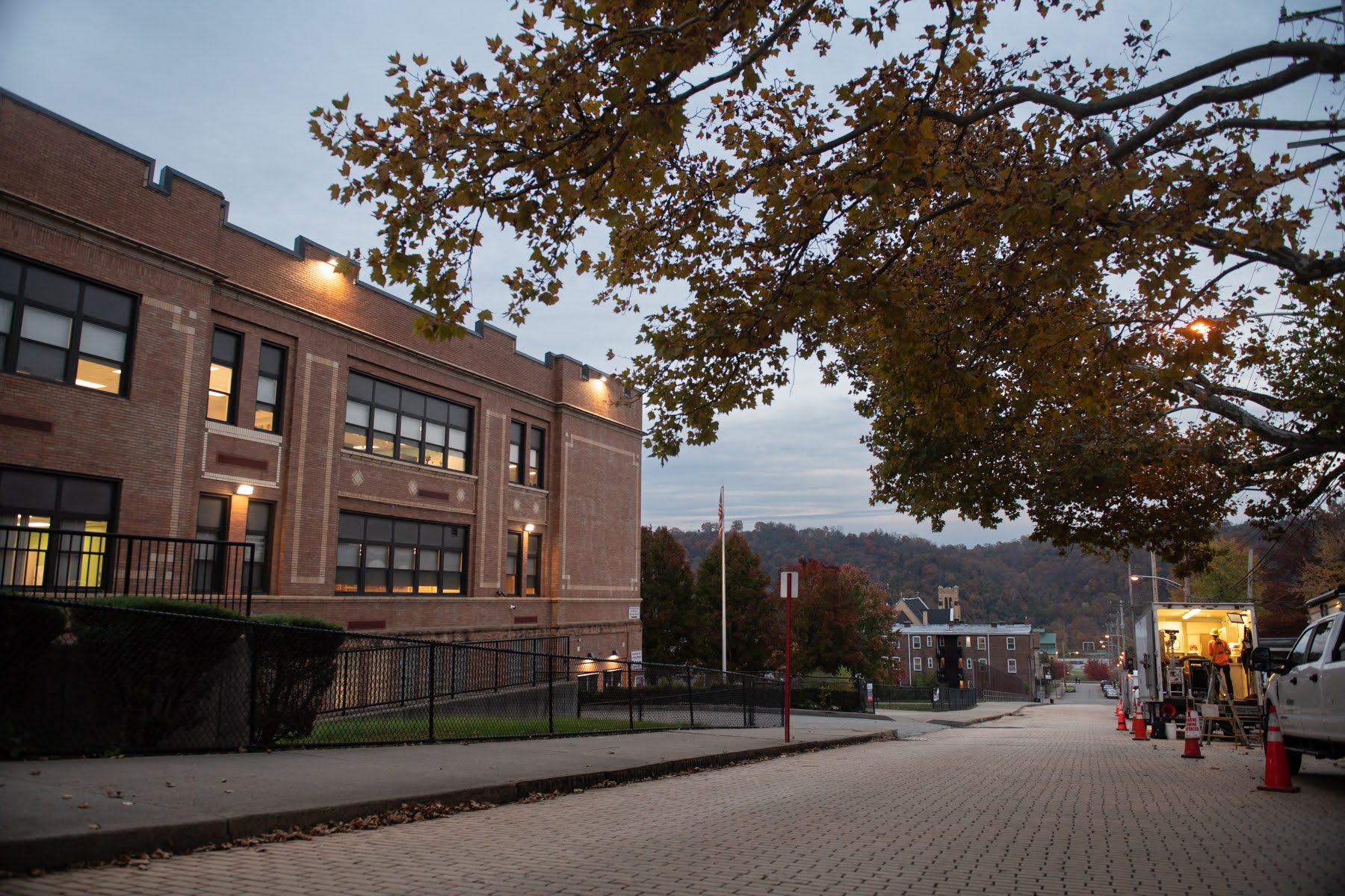 A brick school building and library on a quiet, tree-lined street at dusk, with lights glowing inside and scattered traffic cones along the sidewalk.