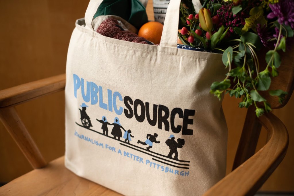A Publicsource tote bag sitting on a chair, filled with a bouquet of flowers, an umbrella, and fruit. The tote reads PUBLICSOURCE: Journalism for a better Pittsburgh and shows images of a person in a wheelchair, a person in a headscarf holding the hands of two children, a person running, and a person carrying a stack of books.