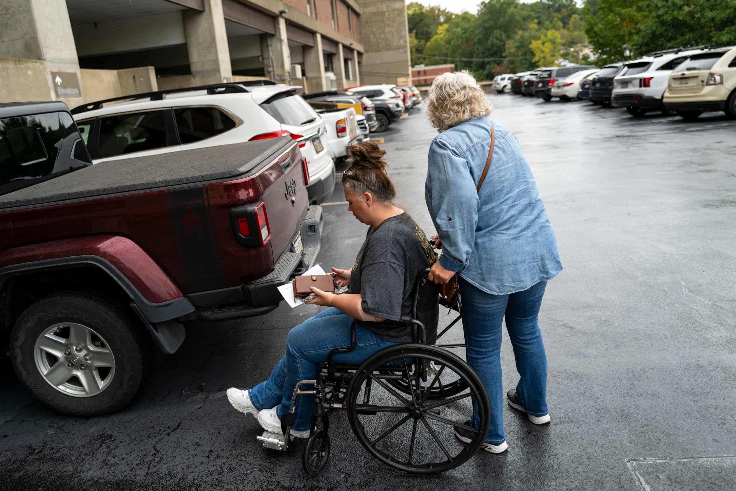 A woman in a wheelchair is pushed by another woman in a parking lot.