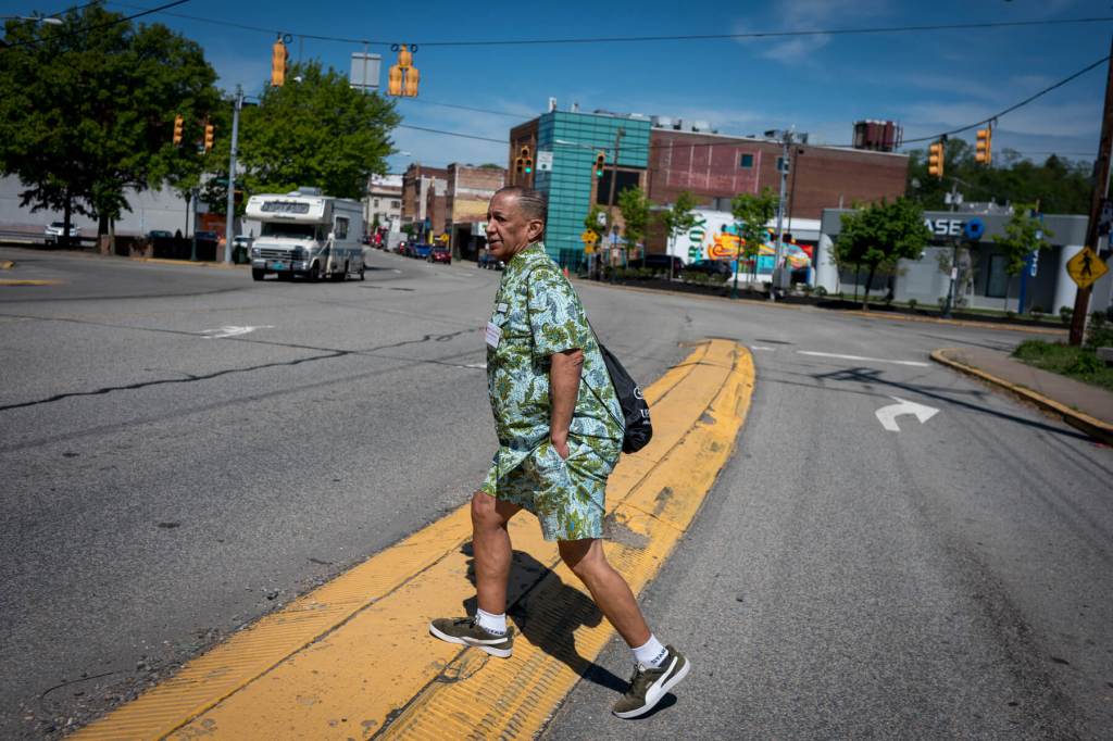 A man wearing a green patterned outfit and a black shoulder bag crosses the street on a sunny day, with buildings and a truck in the background.