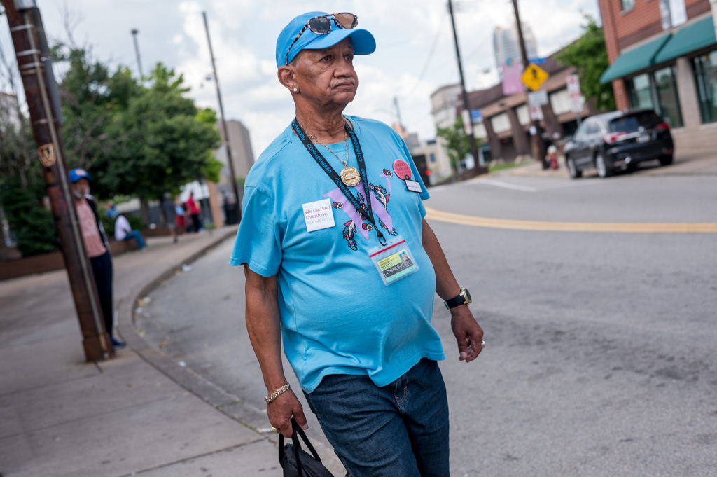 An elderly man wearing a blue T-shirt and cap walks along an urban street carrying a black bag, with buildings and cars in the background.