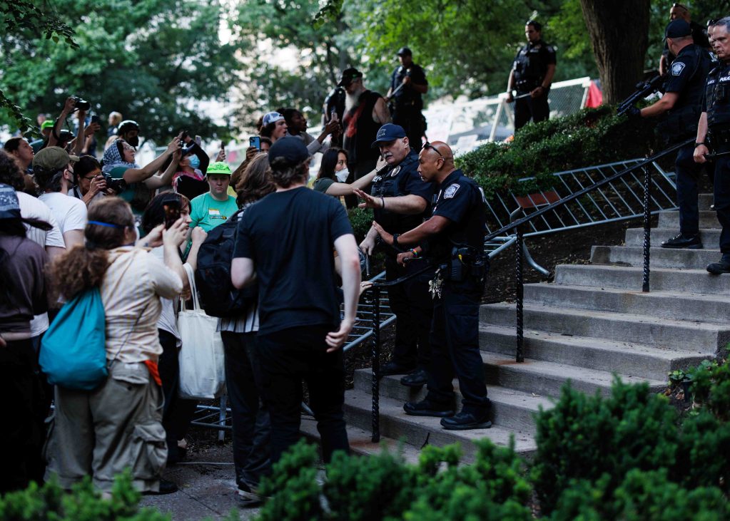 A group of police officers blocks a staircase while facing a crowd of people. Some individuals in the crowd are taking photos and holding signs. Trees and greenery are visible in the background.