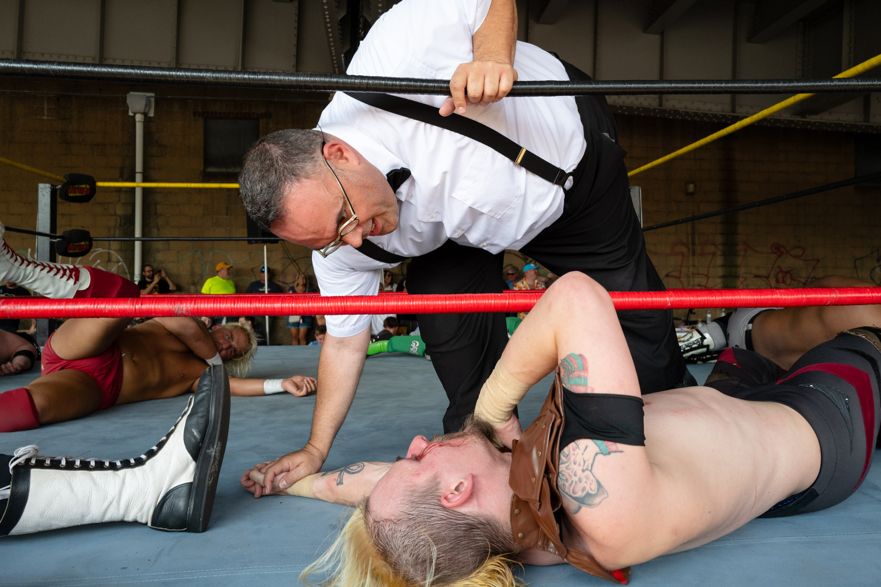 A man in a white shirt and suspenders checks on a wrestler lying on the ring floor. Another wrestler lies in the background.
