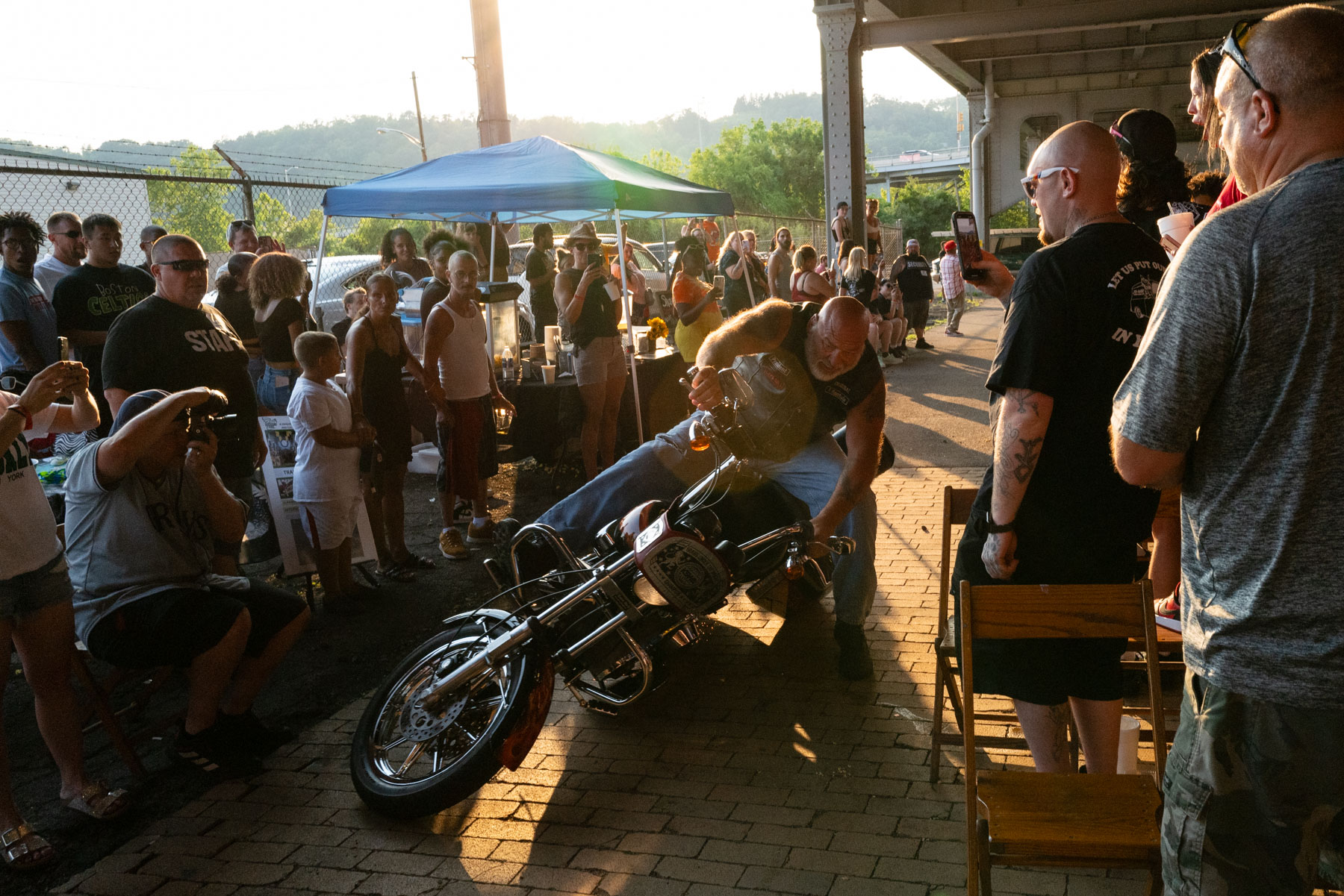 A crowd watches as a person appears to lose control of a motorcycle on a sunny day. The event takes place outdoors near a tent and other spectators.