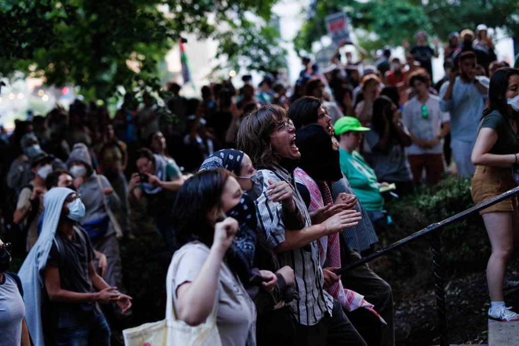 A large crowd of people gathered outdoors, many are shouting and some wearing masks. Trees and foliage are visible in the background.