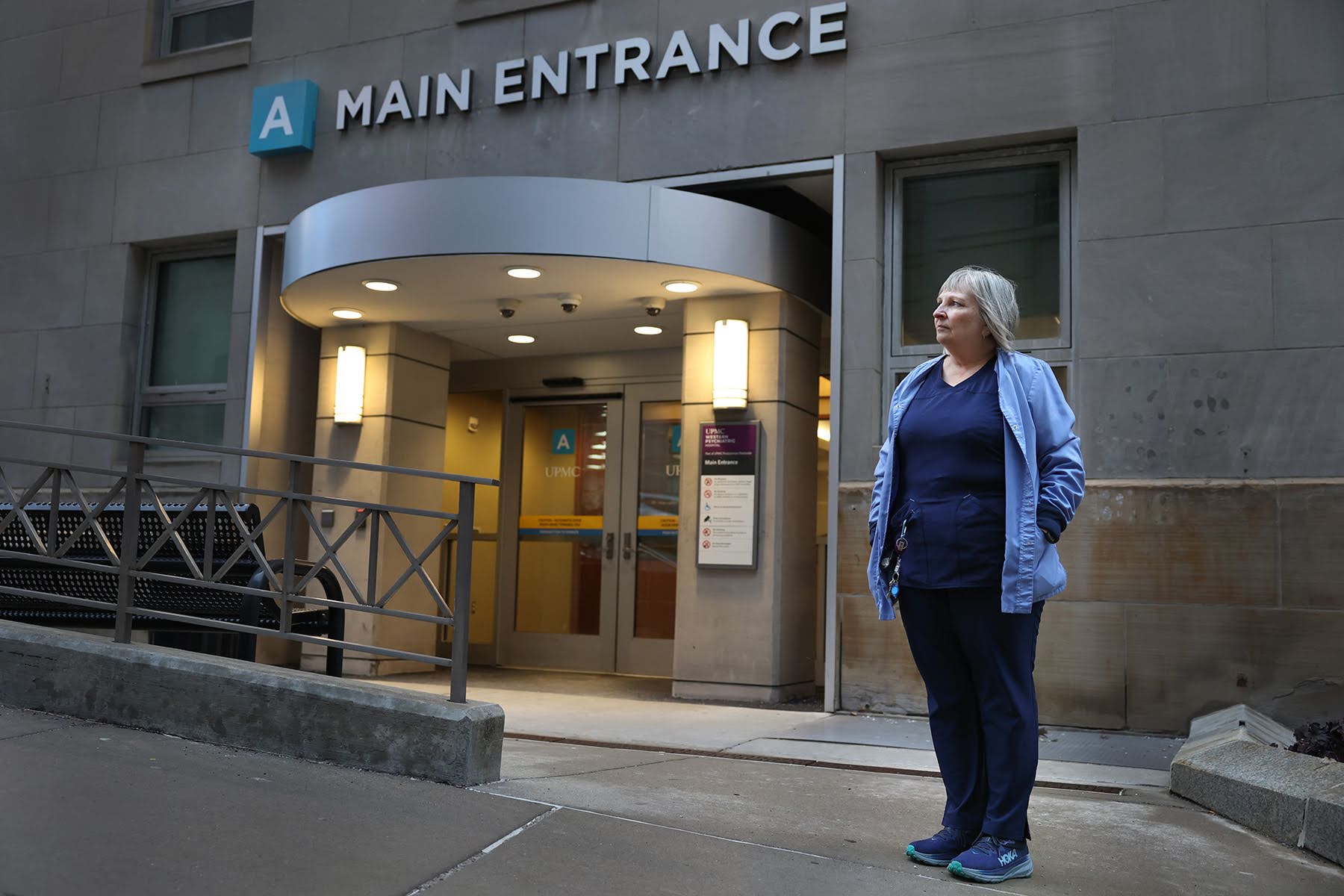 A person in scrubs stands outside a hospital's main entrance.