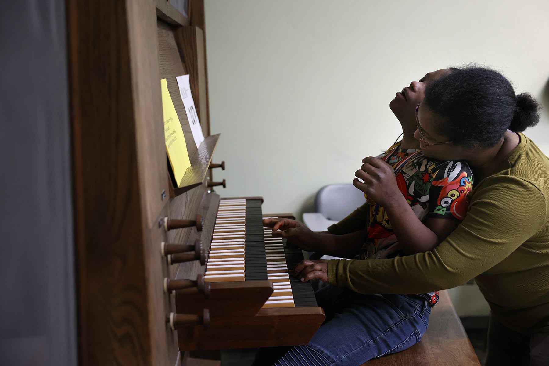 Two people sit closely on a bench playing a wooden organ. One person is resting their back on the other's chest while reaching for the keys. Papers are placed on the organ stand.