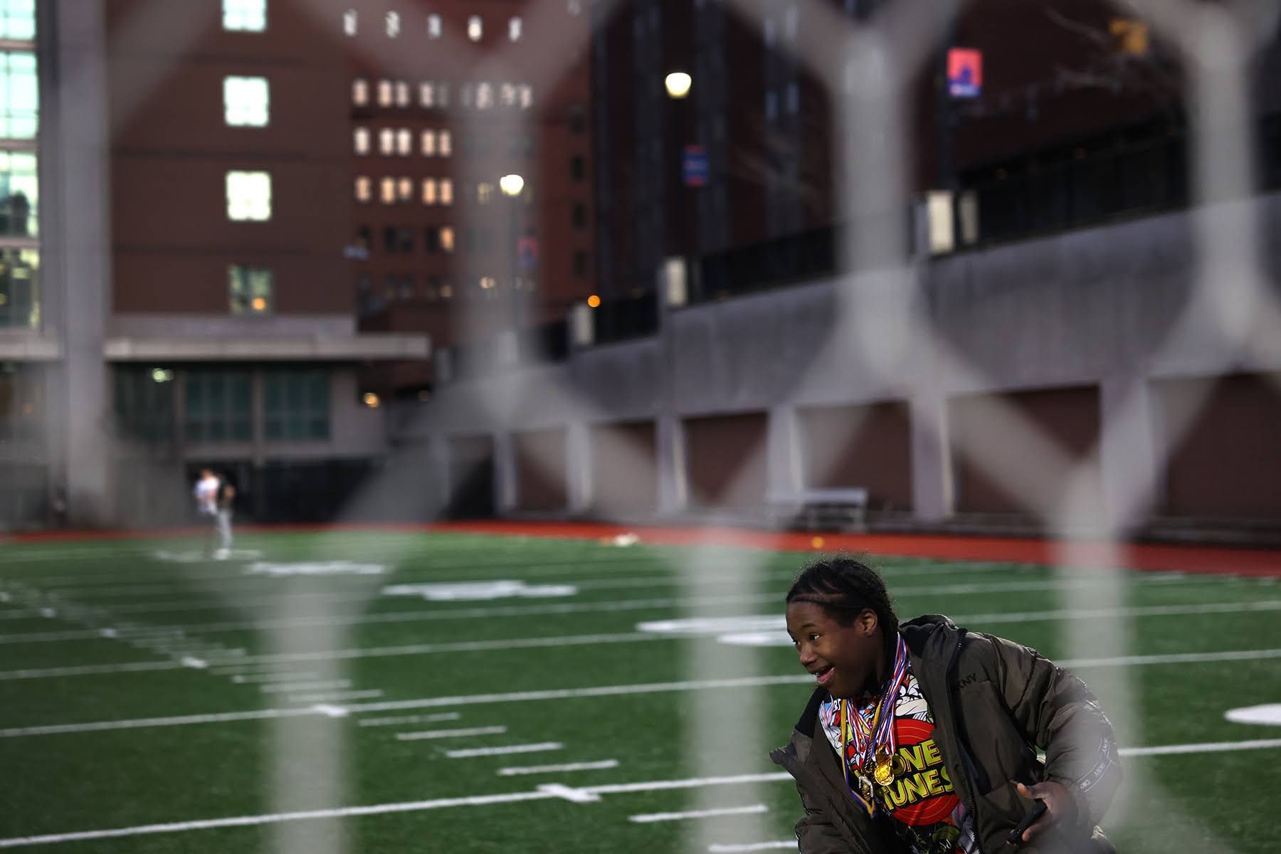 Person with a colorful shirt and jacket stands on a sports field near a goal, with buildings and another person in the background, viewed through a net.