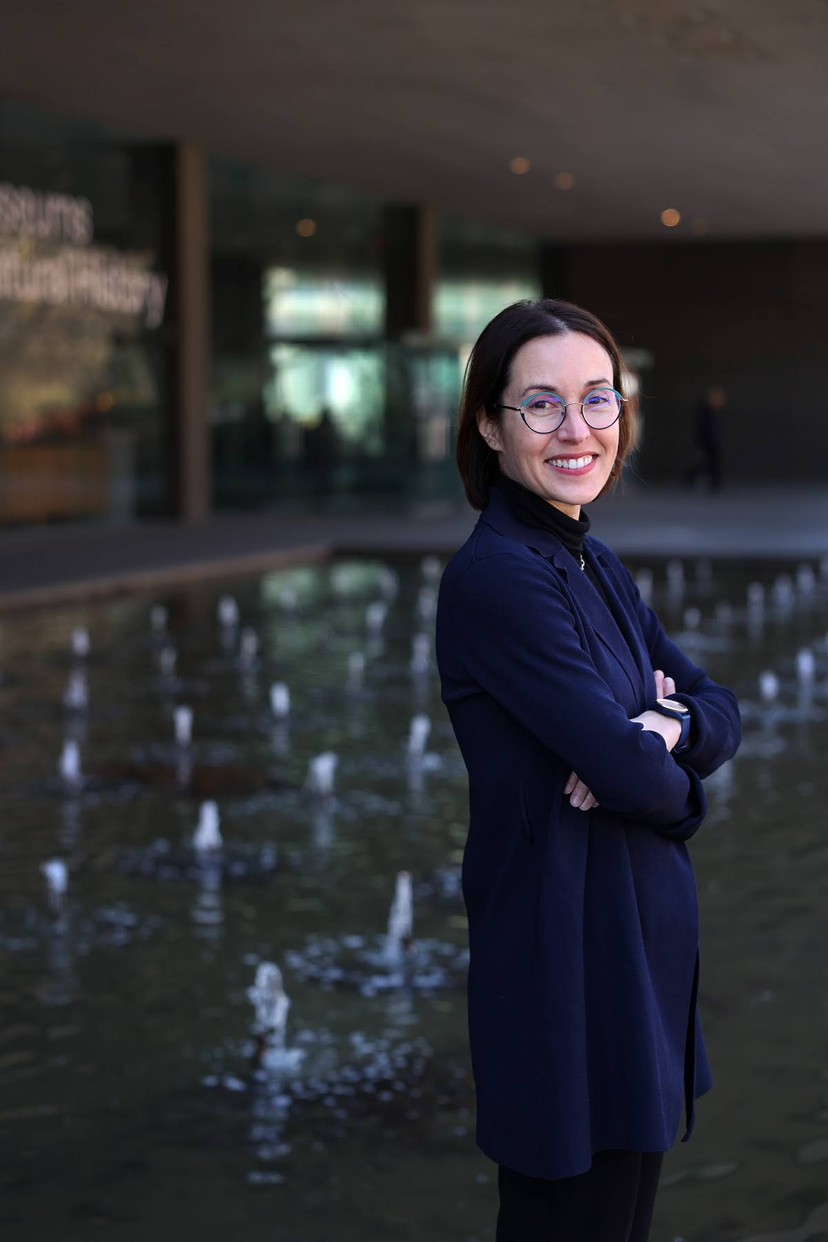 A woman wearing glasses and a dark jacket stands smiling, arms crossed, in front of a modern building and small water fountains.