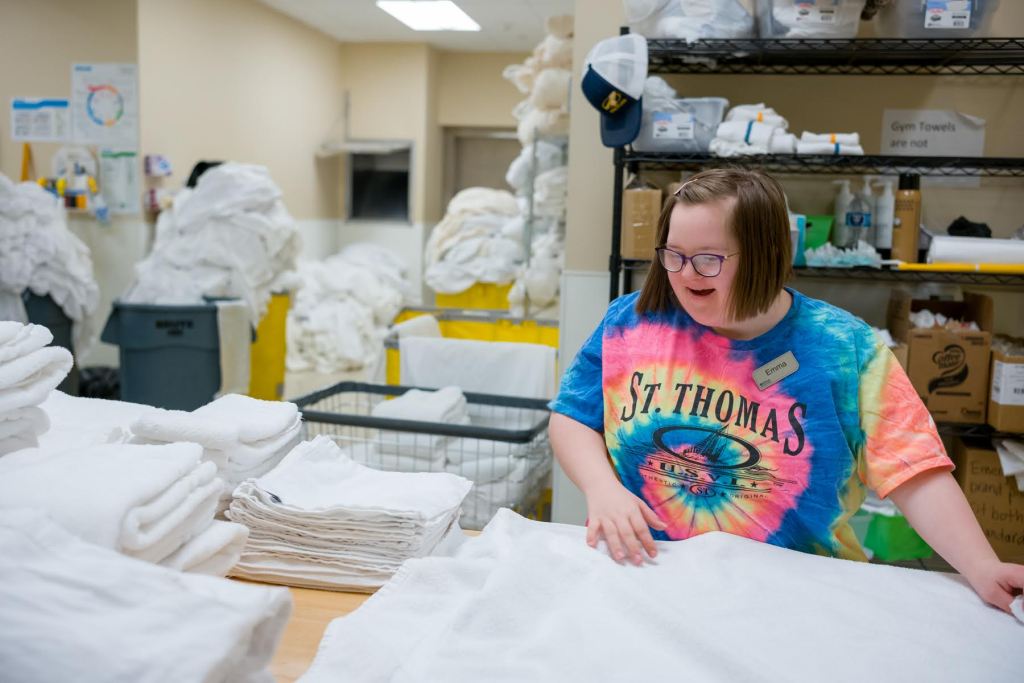 A person folds white towels in a laundry room, wearing a tie-dye T-shirt with the words 