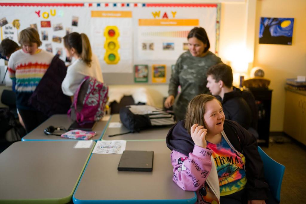 Students in a classroom with colorful posters on the wall. A girl in the foreground smiles, wearing bright clothes. Desks have laptops and backpacks.
