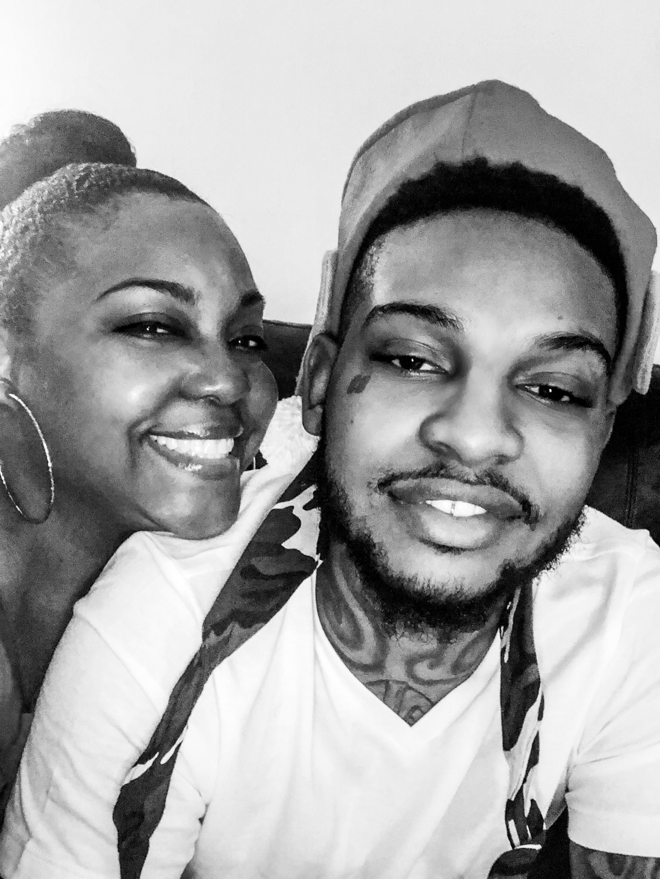 A woman and a man smile for a close-up selfie. The woman has hoop earrings and the man has tattoos and wears a cap turned backwards. The photo is in black and white.