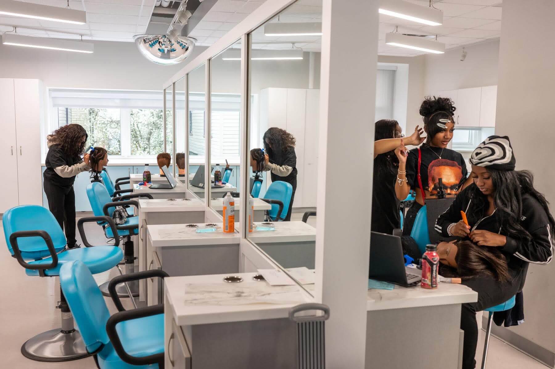 Three girls work on wigs at salon stations with mirrors and blue chairs in a modern, well-lit hair salon or cosmetology classroom.