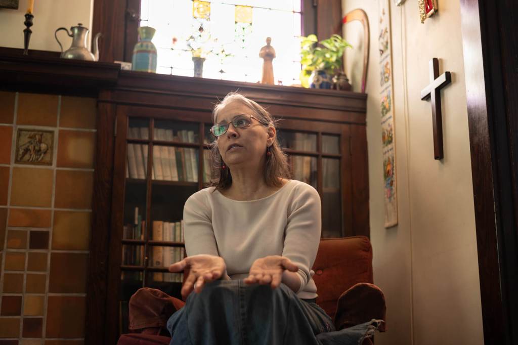 A woman wearing glasses and a white shirt sits on an armchair in her home. A large Christian cross hangs on the wall above her.