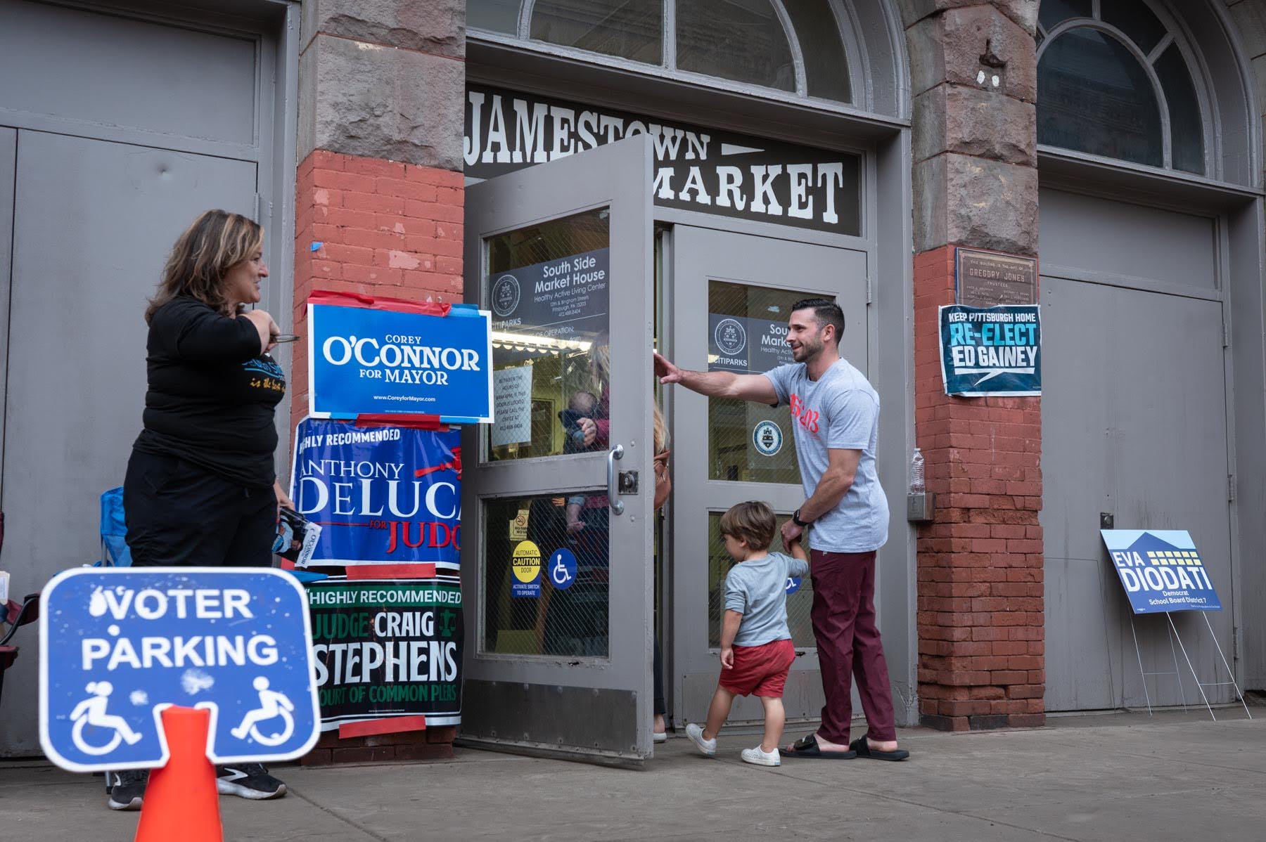 A man holds open the door to Jamestown Market for a child while political campaign signs are displayed outside, including a 