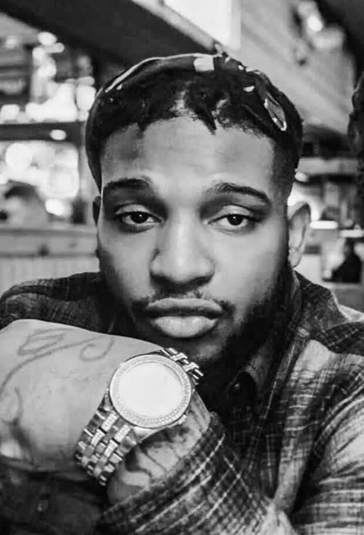 A man with short hair and facial hair, wearing a patterned shirt and a large wristwatch, looks directly at the camera in a black and white indoor setting.