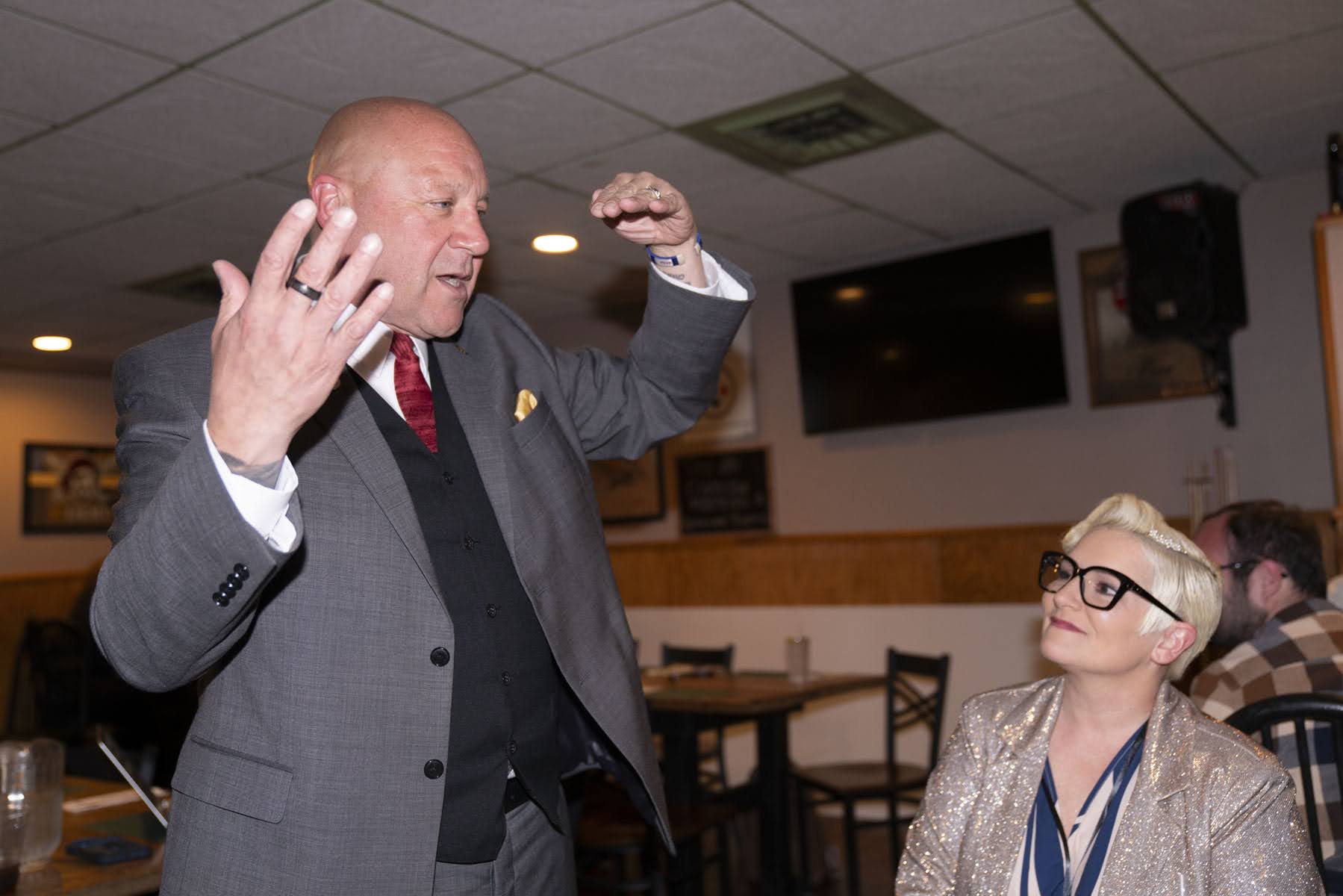 A man in a suit gestures animatedly while standing, as a woman with short blond hair and glasses sits and looks up at him in a restaurant setting.