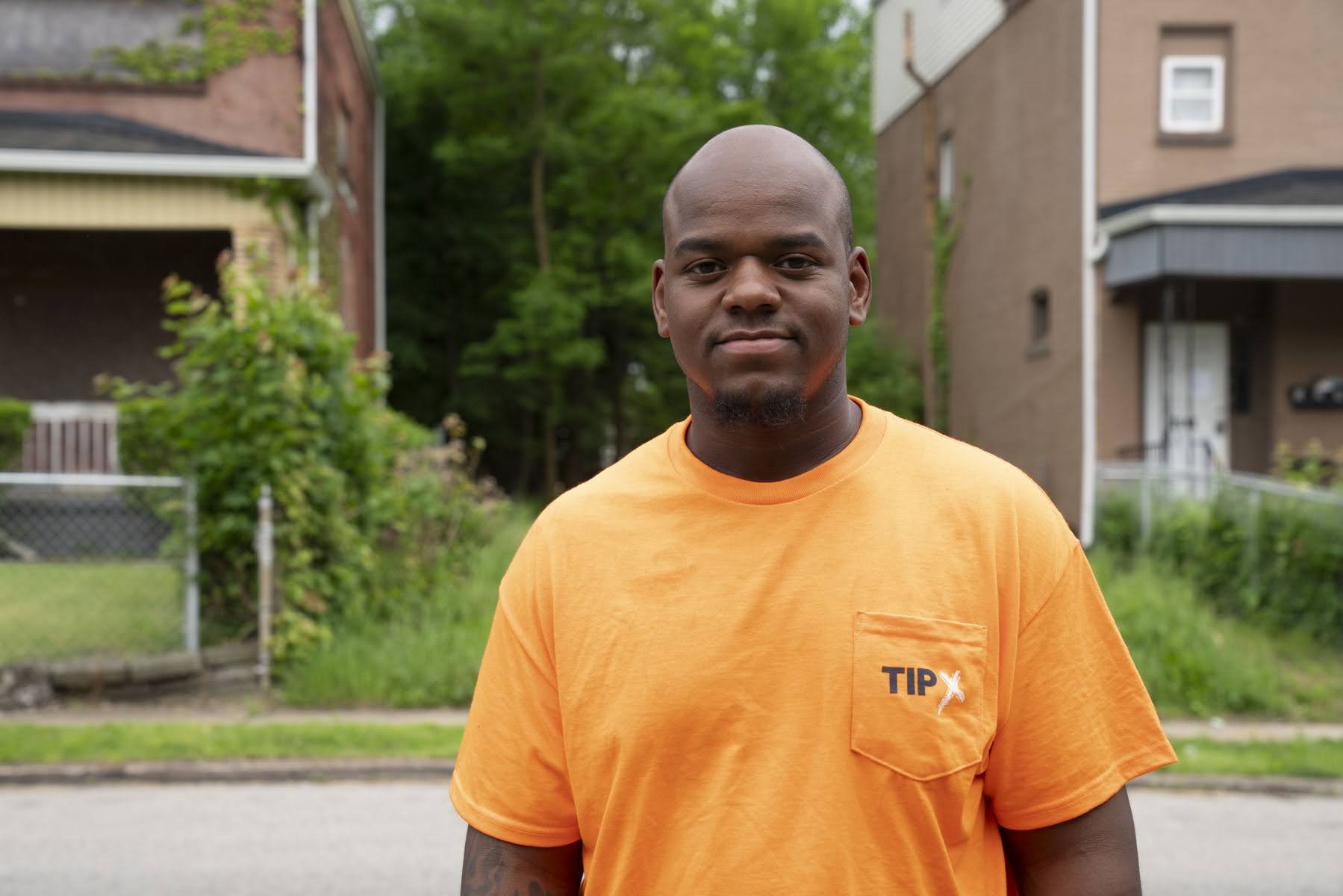 A man wearing an orange T-shirt stands outside on a residential street with houses and greenery in the background.