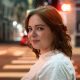 A person with shoulder-length brown hair and a white shirt stands on a city street at night, looking over their shoulder at the camera.