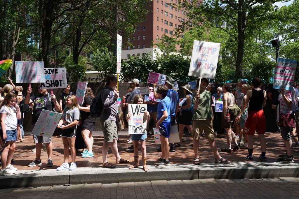 A diverse group of protestors gathers outside UPMC's corporate offices in Downtown Pittsburgh, holding signs advocating for transgender rights and opposing the recent cut of gender-affirming care for patients under 19.
