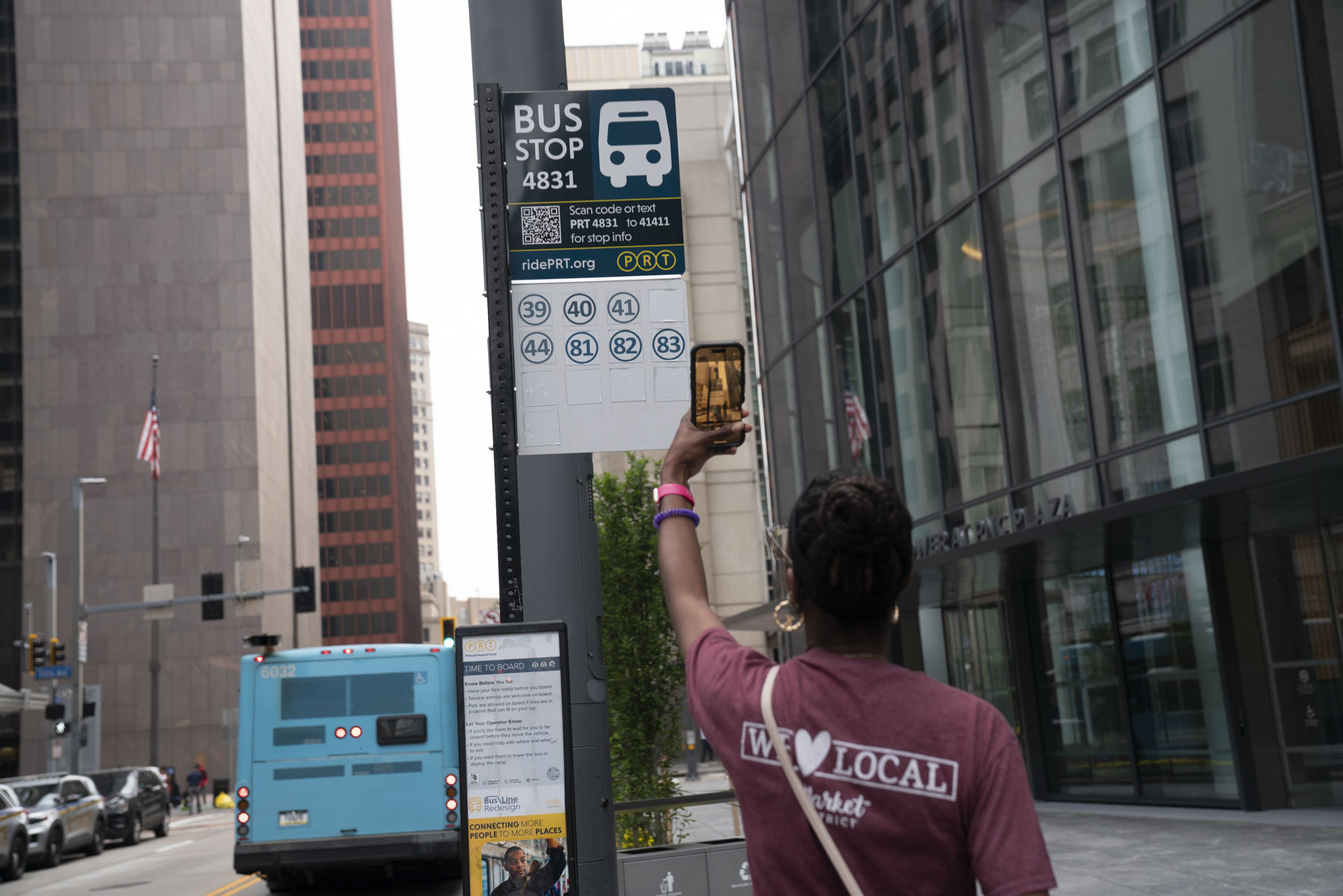 Person in a maroon shirt stands at a city bus stop, and takes a picture of a sign with route numbers, as a blue bus drives down the street amid tall buildings.