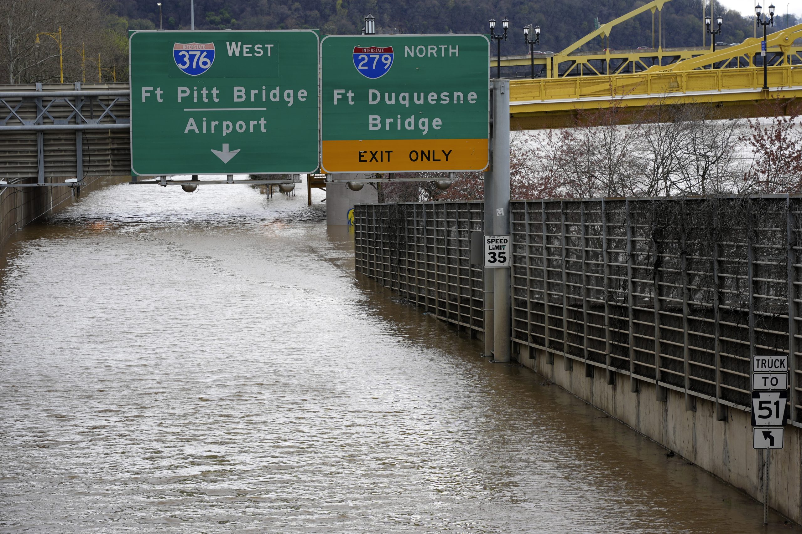 A highway entrance is flooded, submerging lanes under water. Overhead signs indicate directions to I-376 West and I-279 North bridges. A speed limit sign and trees are visible.