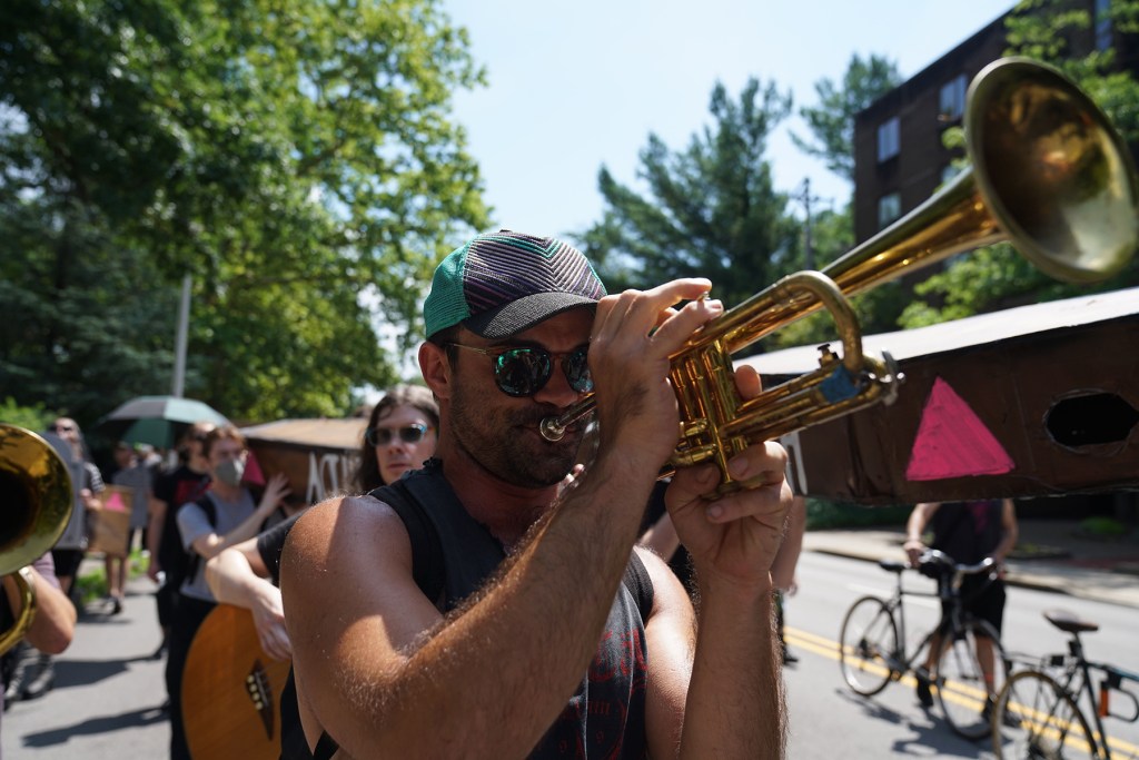 A man in sunglasses and a cap plays a trumpet during an outdoor street event, with other people, bicycles and trees in the background.