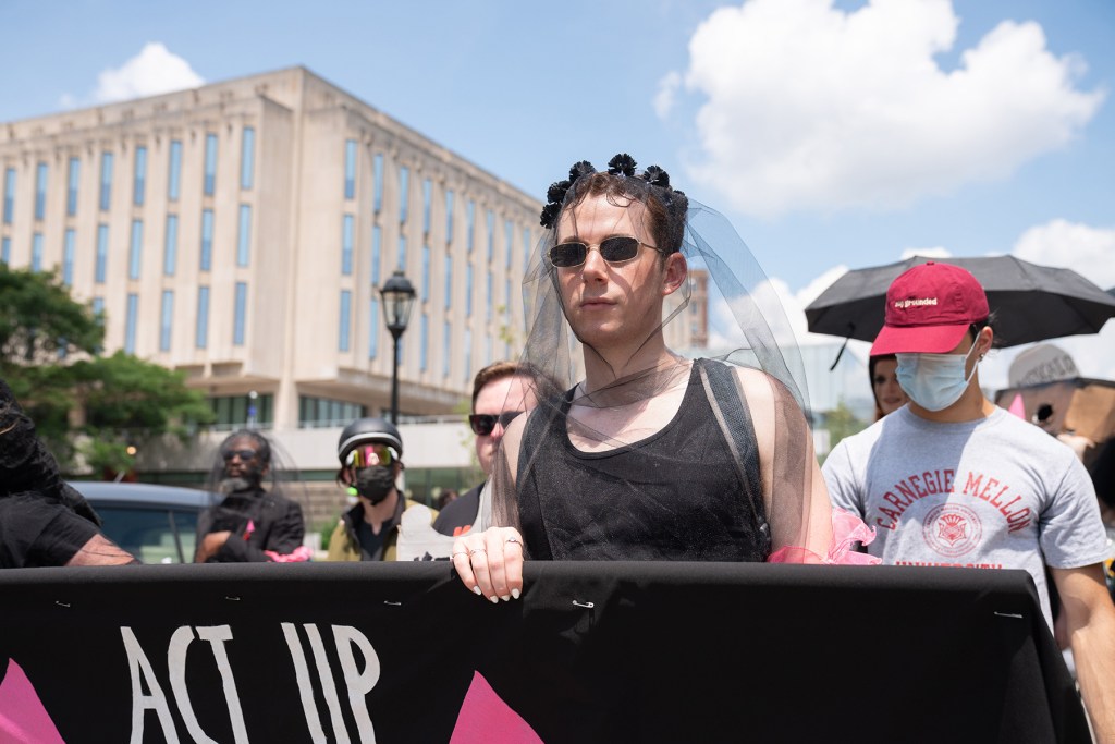 A person in a black outfit and veil stands at the front of a protest holding an ACT UP banner. Other people are behind, some wearing masks, near a large building on a sunny day.