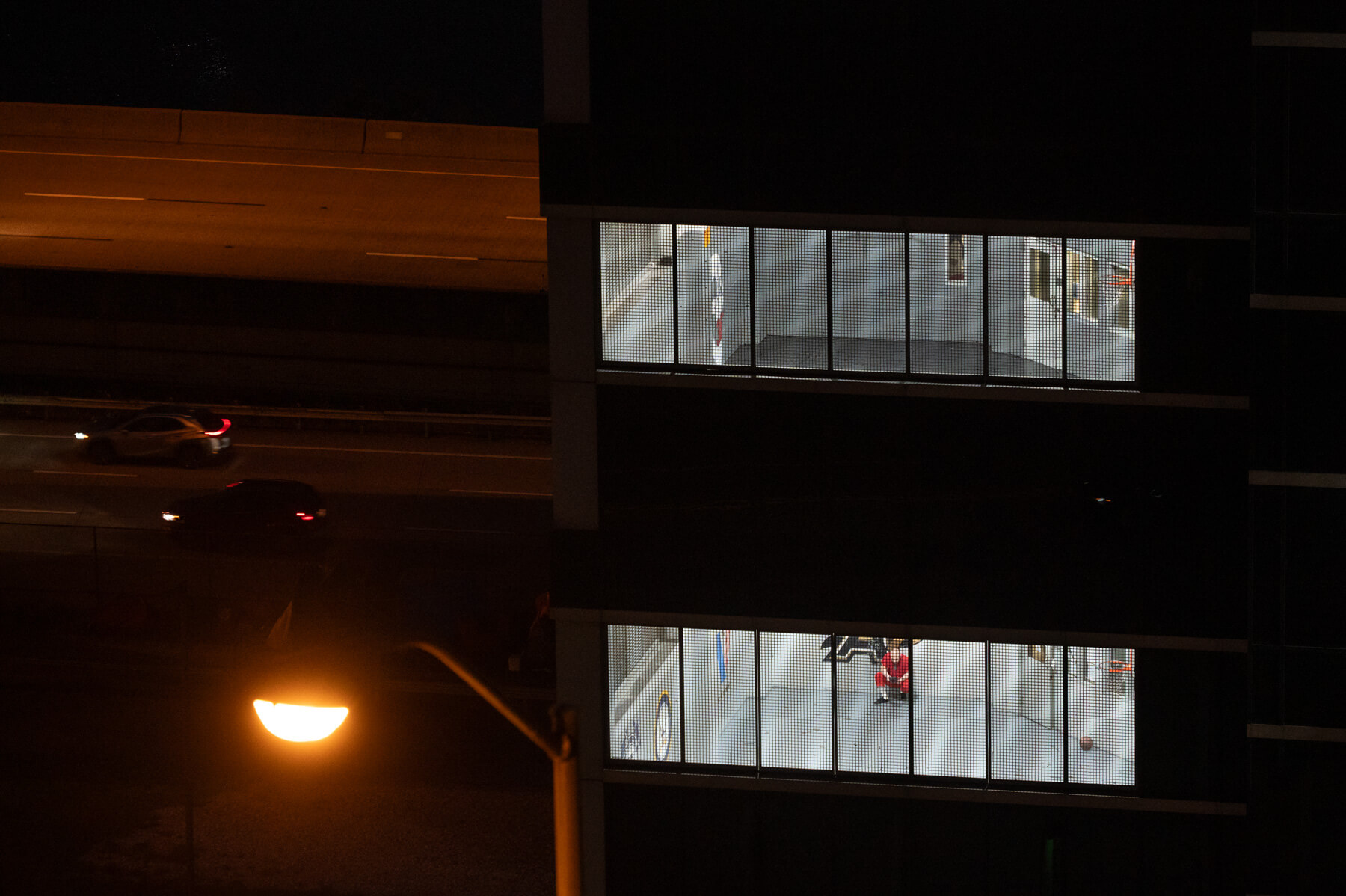 A jail building with two brightly lit rooms and visible wall art is seen at night, with cars driving on a nearby highway and a streetlamp in the foreground. A person sits in the empty room with fencing over the windows.