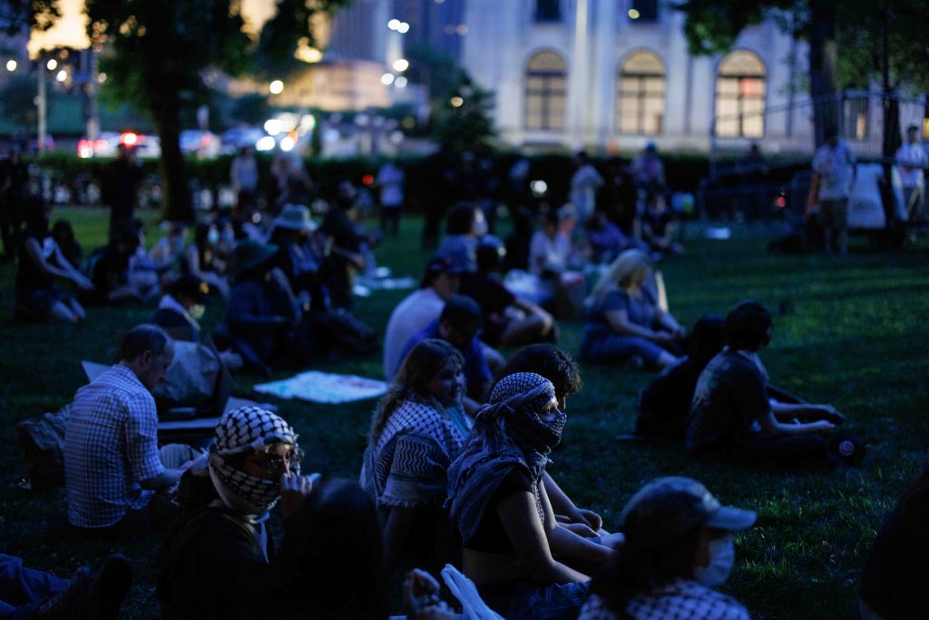 A group of people sit on the grass in a park at dusk, some wearing masks and patterned scarves, with buildings and city lights visible in the background.