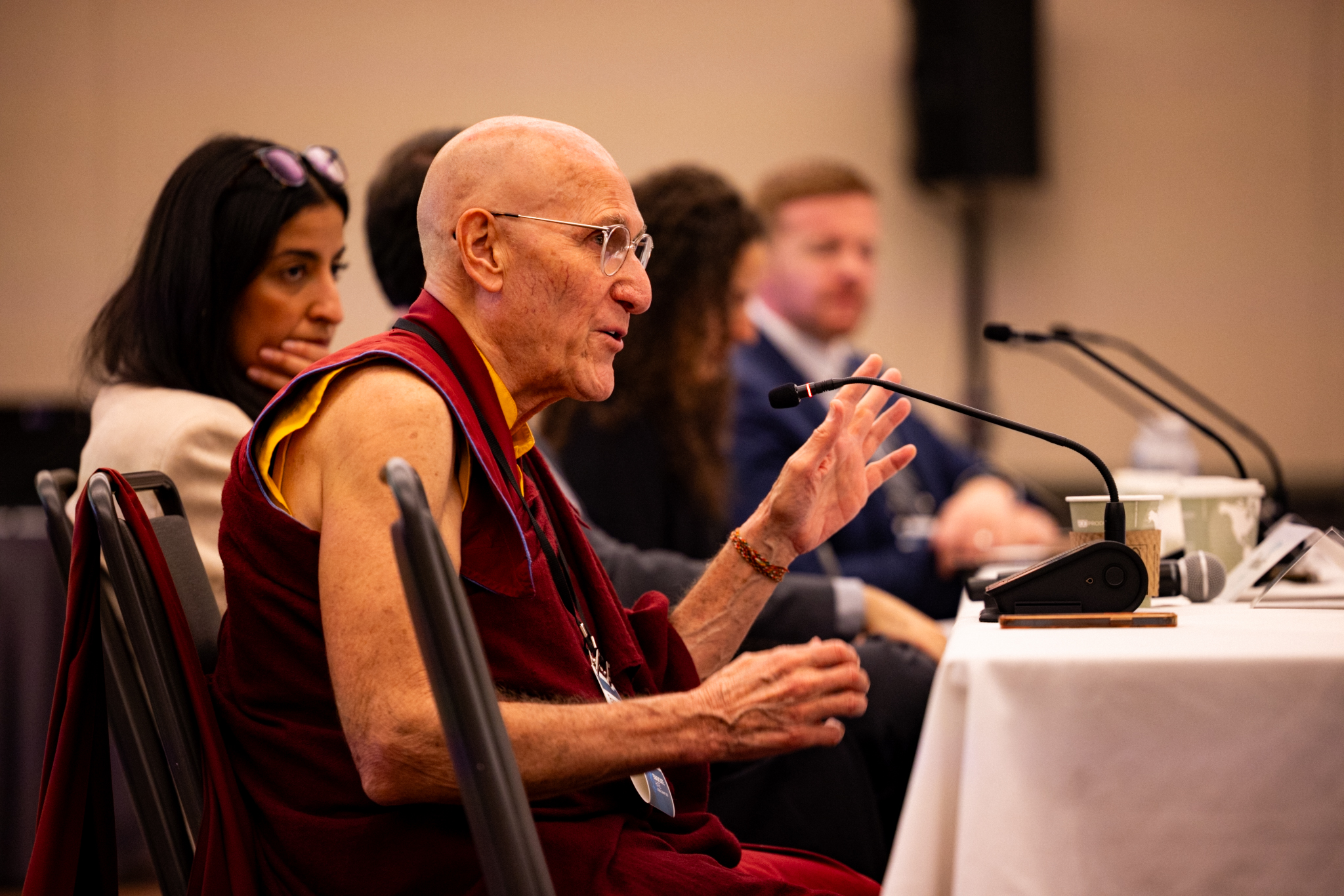 A man in maroon robes speaks into a microphone at a panel discussion, seated with three other people at a conference table.