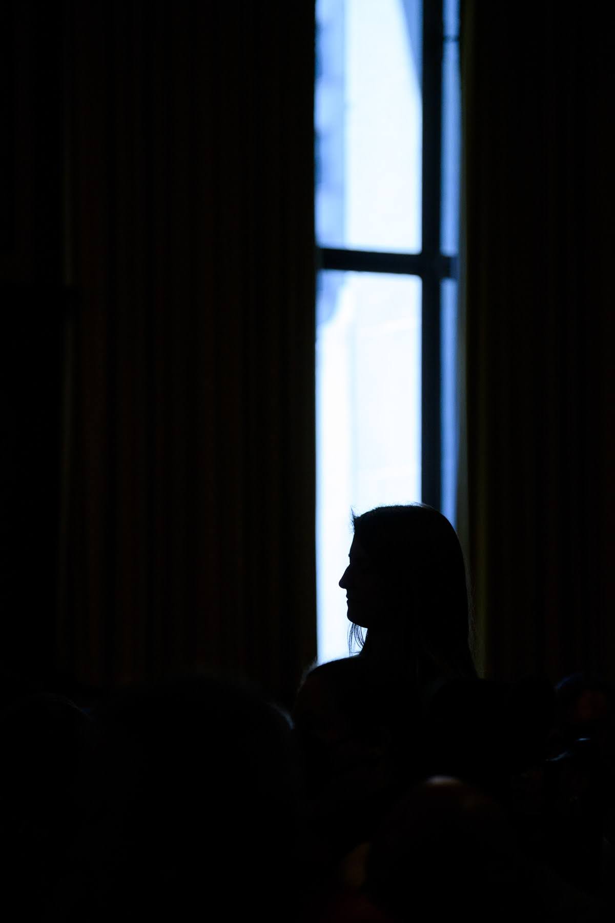 A silhouette of a person with long hair stands in front of a tall window with drawn curtains, illuminated by daylight from outside.