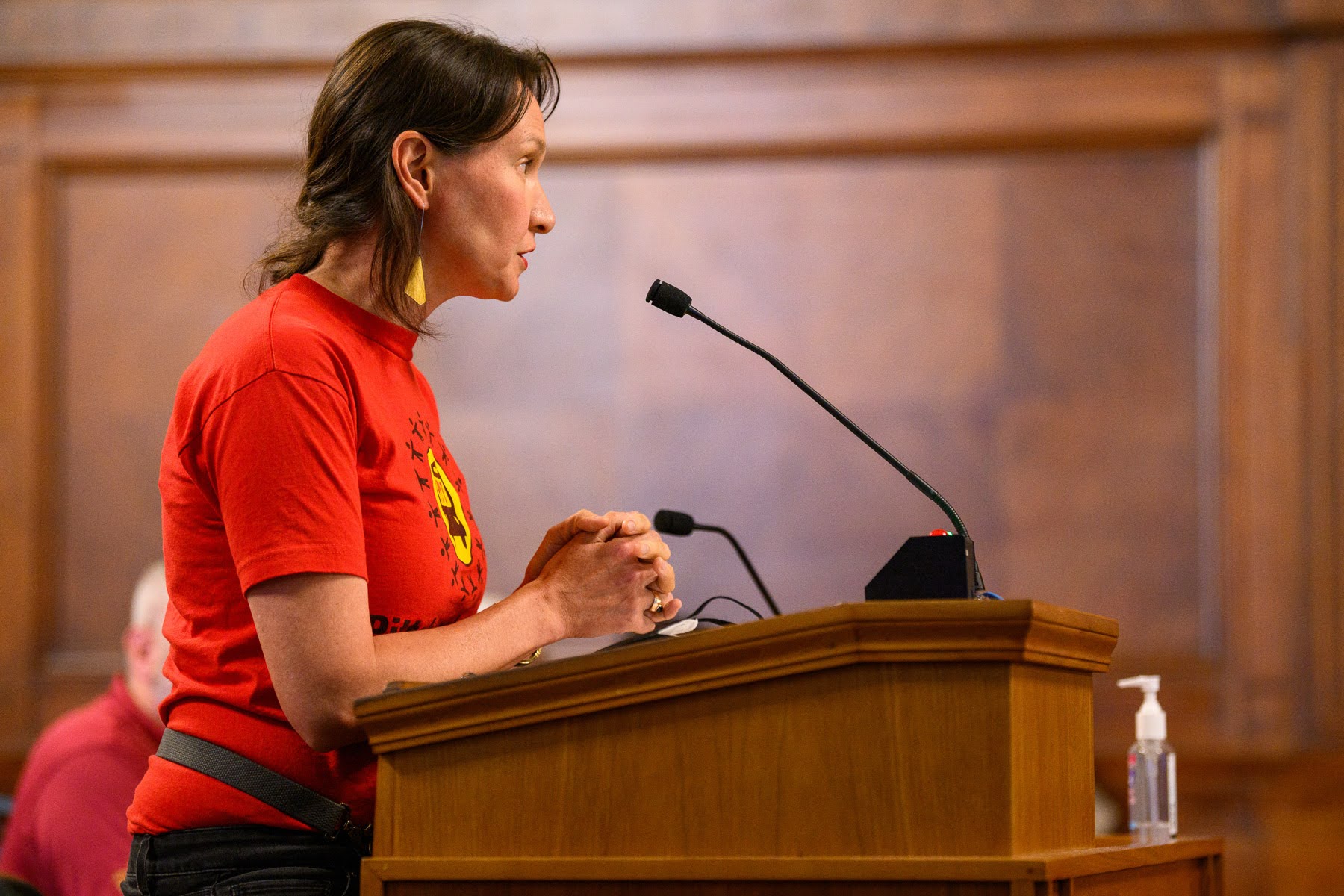 A person in a red t-shirt speaks at a podium with a microphone in a wood-paneled room; a bottle of hand sanitizer is visible on the podium.