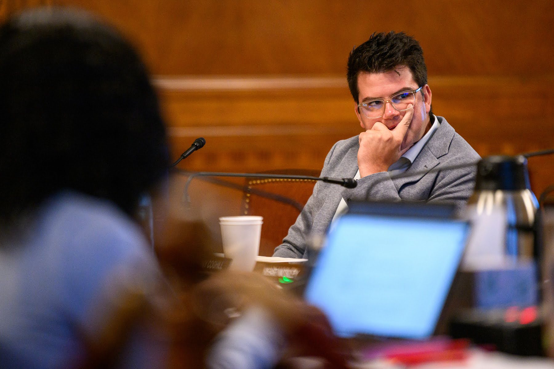A person in a gray jacket sits at a conference table, resting their chin on their hand and looking thoughtful, with laptops and microphones visible in the foreground.