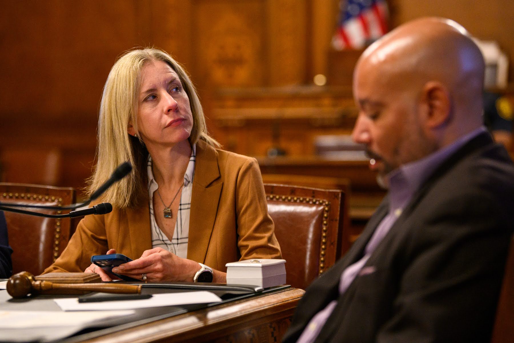 A woman in a brown blazer sits at a desk with papers and a gavel, looking at a man beside her in a courtroom or meeting room setting.