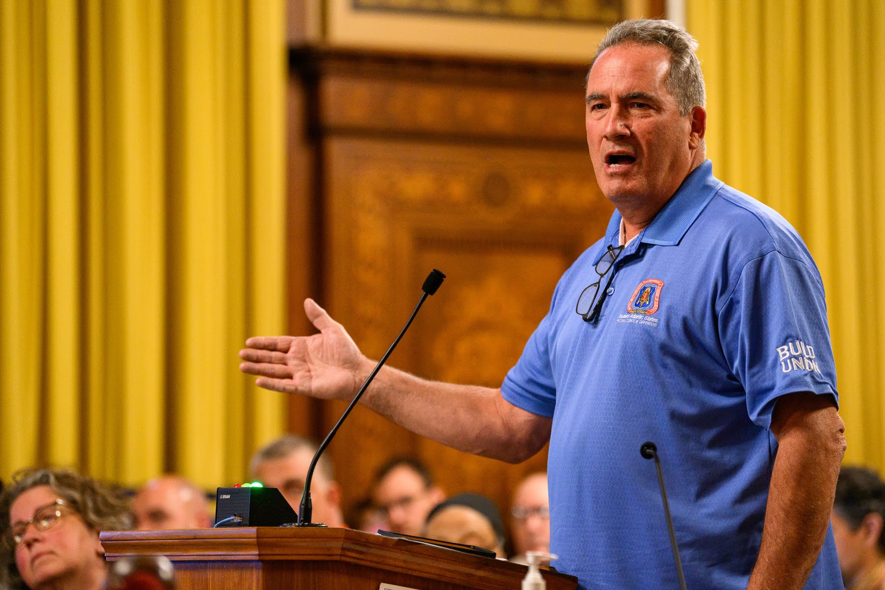 A man in a blue polo shirt speaks at a podium in a formal room, gesturing with one hand. Several people are seated in the background, listening.