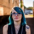 A person with blue hair and glasses stands in an alleyway, wearing a sleeveless top and displaying a sunflower tattoo on their upper arm.