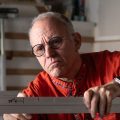 A man wearing glasses and an orange shirt works with metal framing in a workshop filled with tools and equipment.