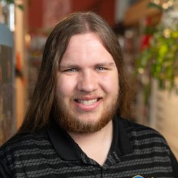 A person with long brown hair and a beard smiles at the camera, wearing a black Pitt Nike polo shirt, standing indoors next to shelves and plants.