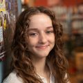 A person with long, curly brown hair and a white shirt leans against a wall in a room with shelves and plants in the background, looking at the camera and smiling slightly.