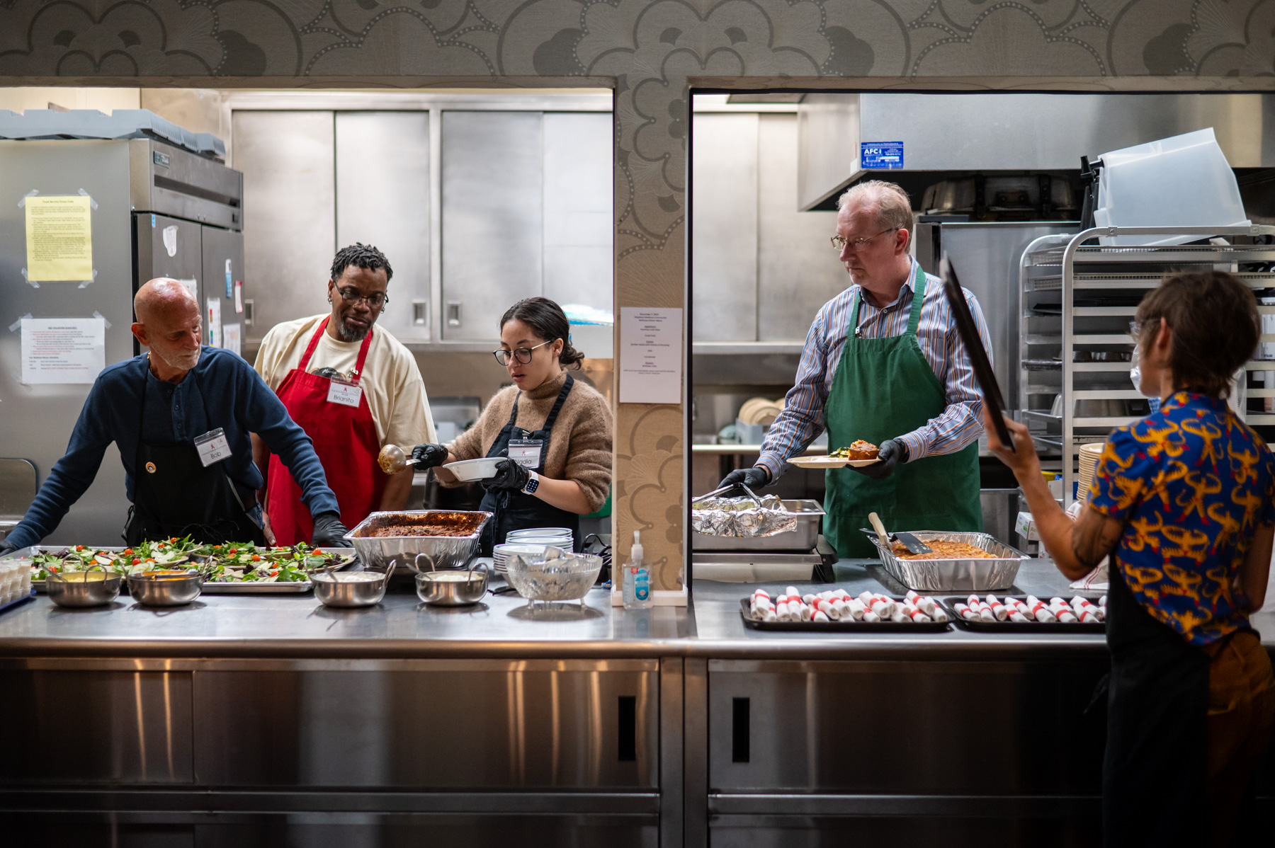 Five people prepare and serve food behind a stainless steel counter in a commercial kitchen. Bowls and trays of various dishes are arranged on the counter.