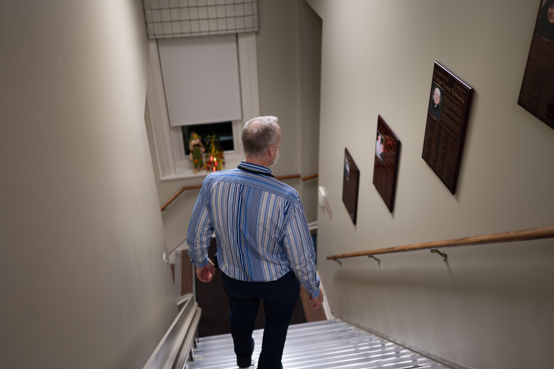 A man with gray hair wearing a striped shirt walks down a staircase with framed plaques on the wall and a window at the landing.