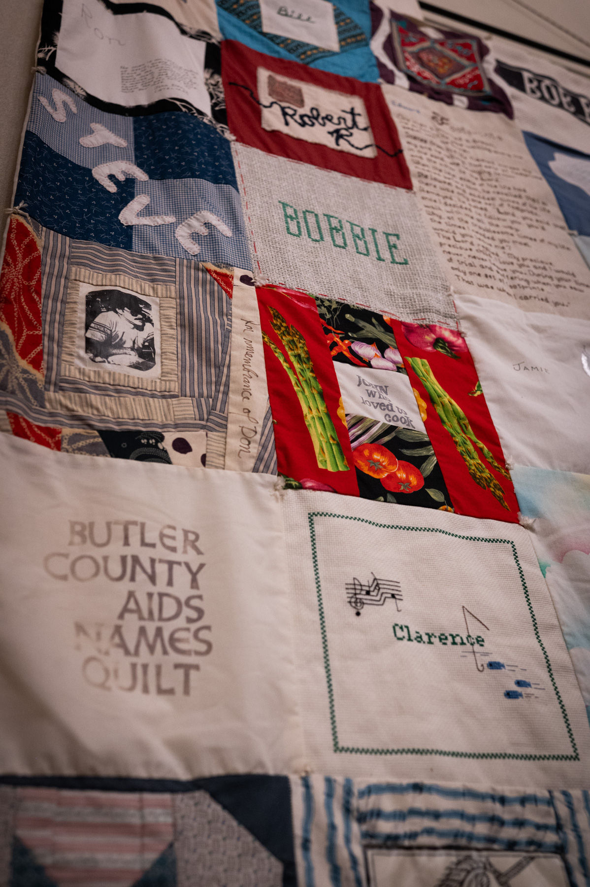 A quilt made of various fabric squares, each with unique designs, names, and text, including “Clarence” and “BUTLER COUNTY AIDS NAMES QUILT.”.