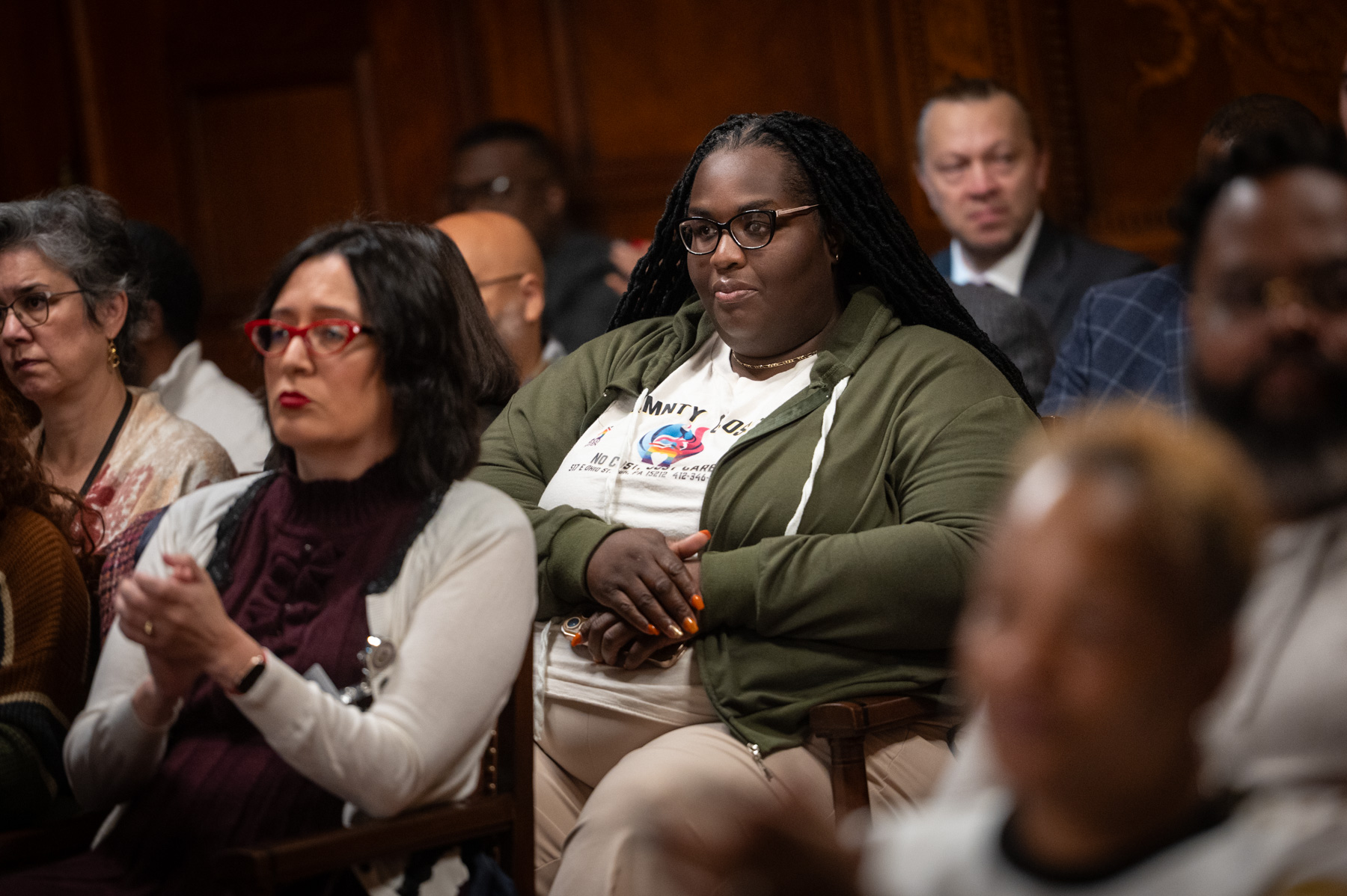 A group of people sit in a wood-paneled room, attentively listening to an event or presentation.