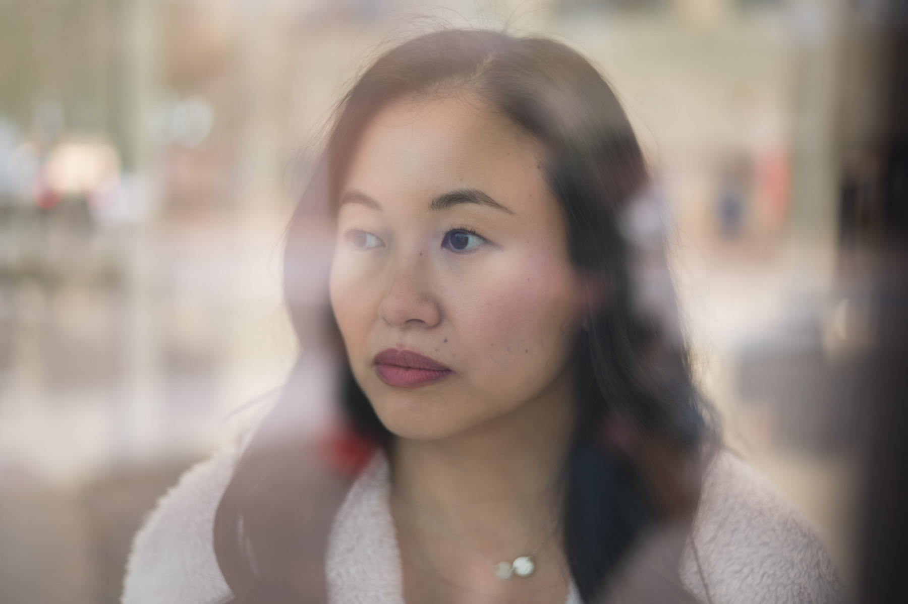 A woman with long dark hair looks pensively out of a window, with reflections and soft natural light visible on the glass.