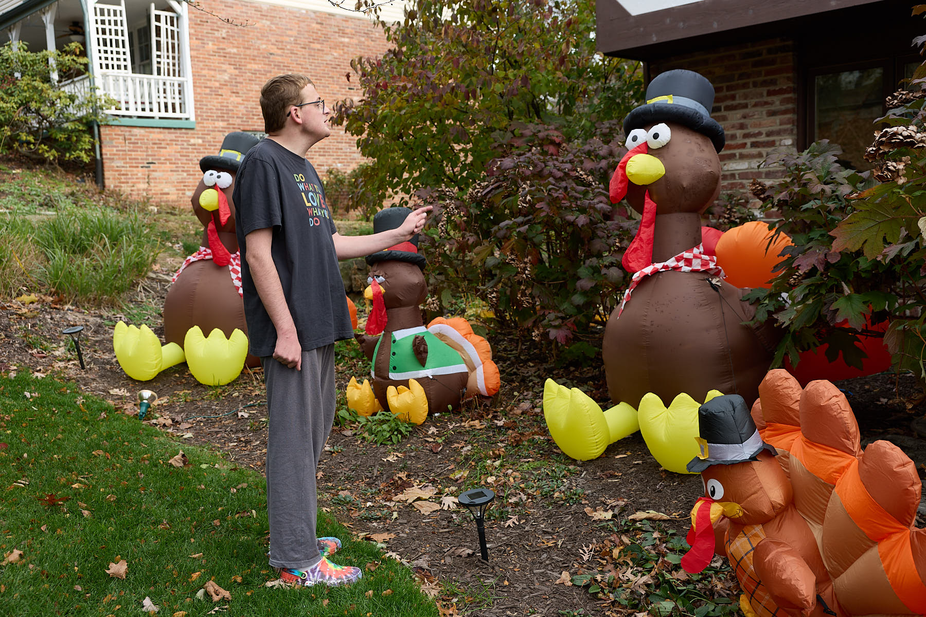 A man in casual clothes stands in a yard decorated with large inflatable turkeys wearing hats and scarves.