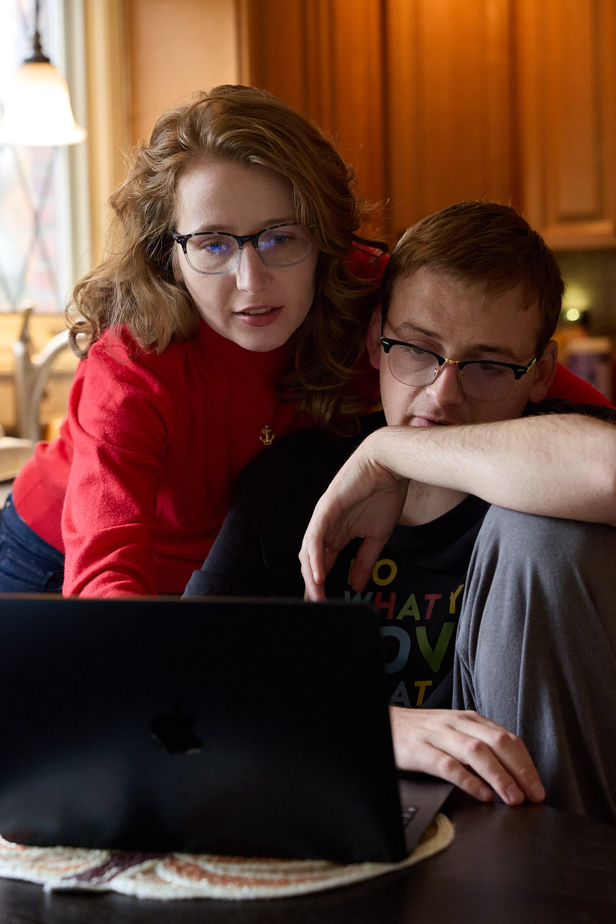 Two people sit closely together in a kitchen, looking at a laptop screen; one points at the display while the other rests an arm on the table.