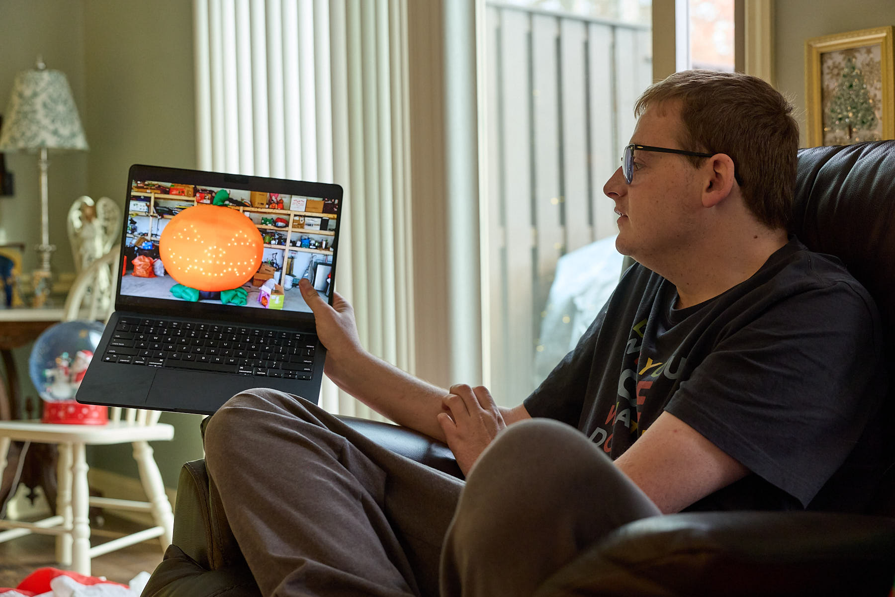 A person sitting in a chair holds a laptop, showing a screen with an orange and multiple video thumbnails in a living room setting.