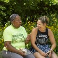 Two women sit on a stone bench in a garden with yellow flowers, engaging in conversation and smiling at each other.