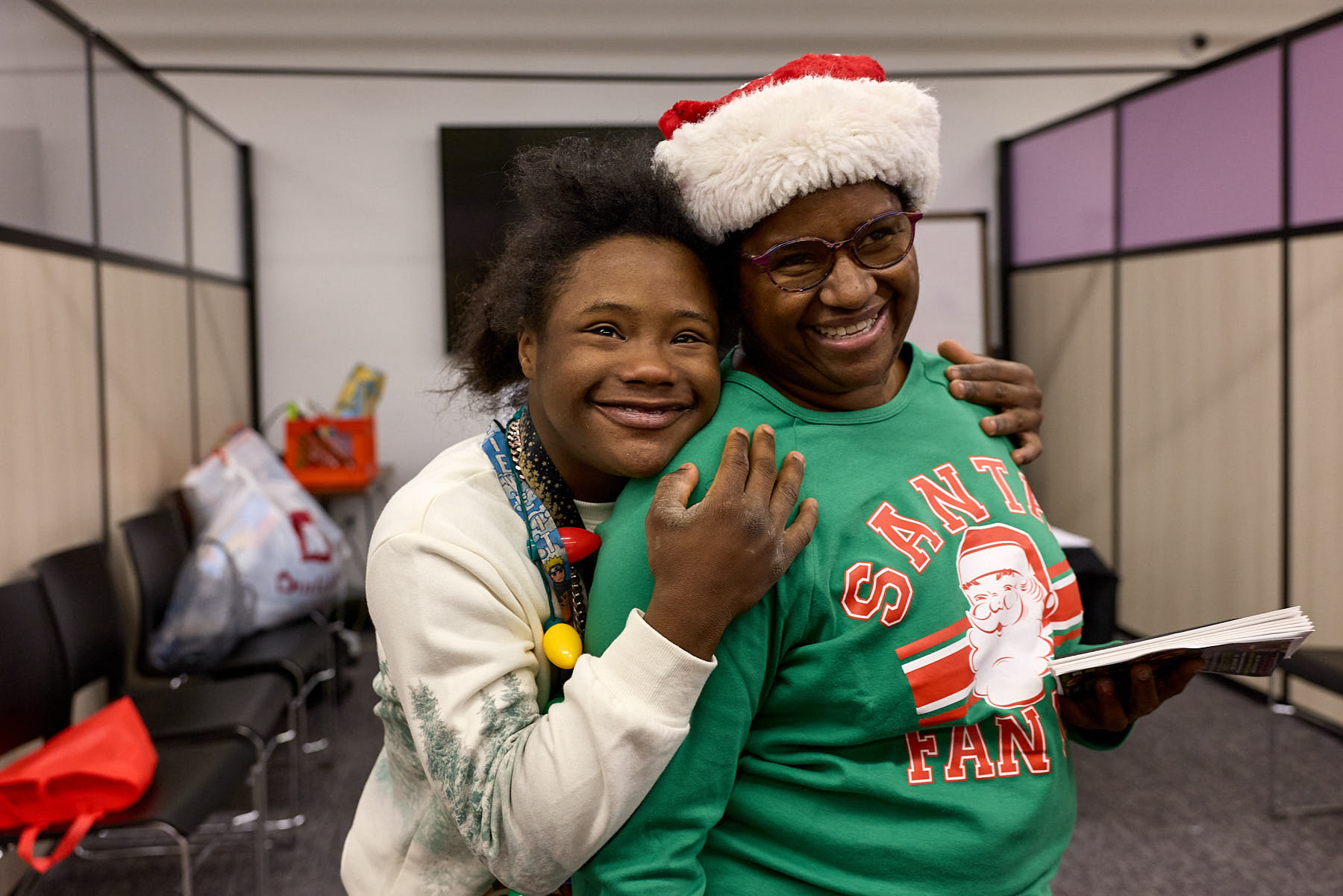 Two people smile and hug in an indoor setting; one wears a Santa hat and festive sweater, the other wears a white sweater with a lanyard.